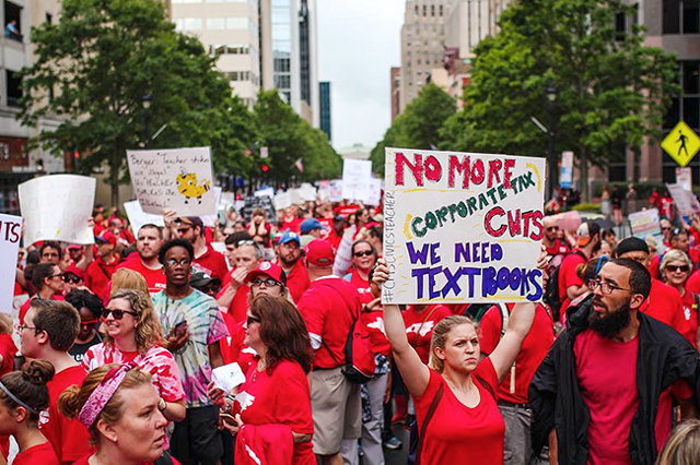 A woman holds up a sign amidst a crowd of others marching in the streets of Raleigh, protesting the corporate tax breaks that companies receive. The sign has the hashtag #CMSCivicsTeacher. (Photo by Danny Timpona)